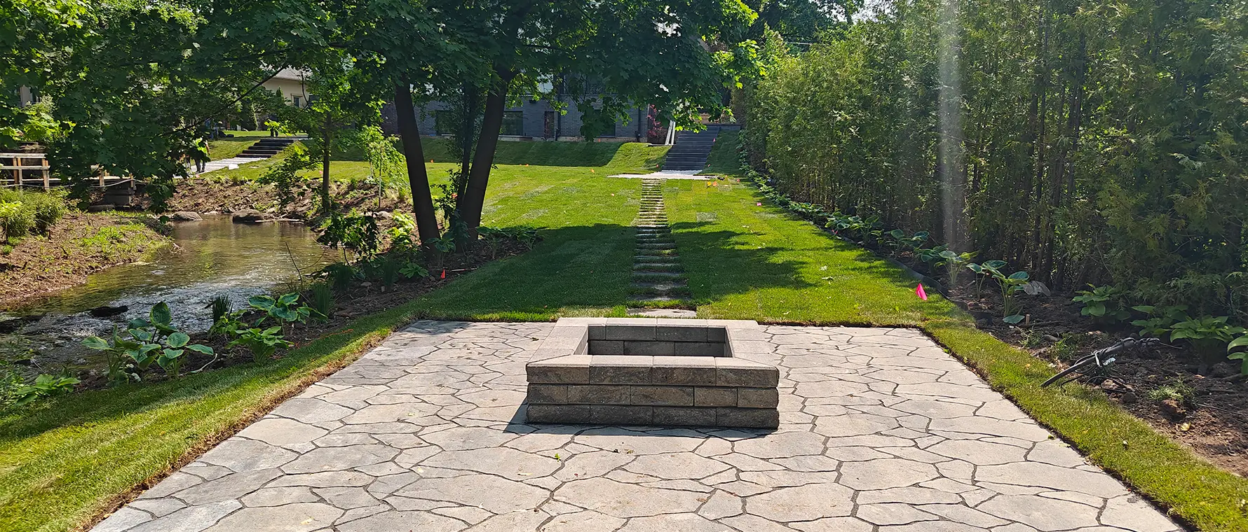 Completed backyard fire pit patio with interlocking stone and landscaped lawn near a natural creek in Mississauga
