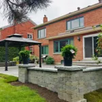Backyard patio with raised stone seating wall, gazebo, and landscaped garden beside a brick home